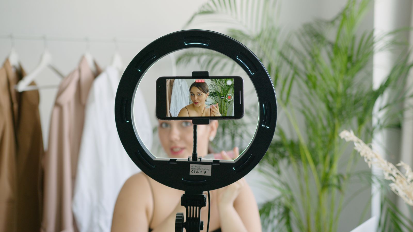 Chủ cửa hàng trực tuyến quay video giới thiệu sản phẩm bằng điện thoại và đèn hỗ trợ trước giá treo quần áo